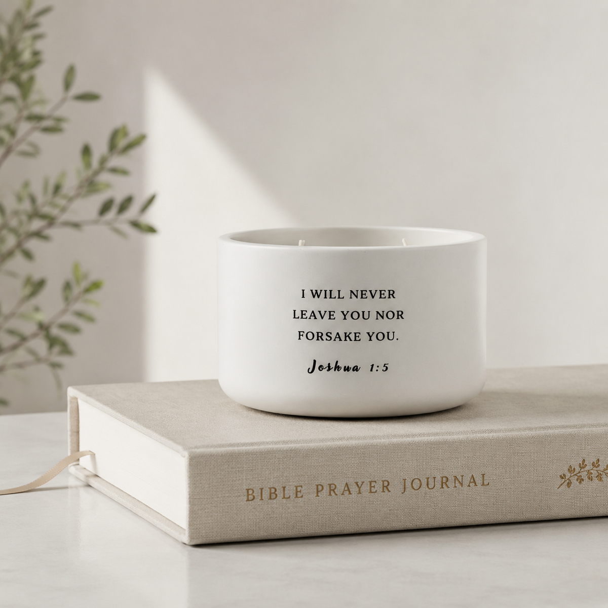 A white ceramic candle jar with black printed text reading 'I WILL NEVER LEAVE YOU NOR FORSAKE YOU. Joshua 1:5' sits on a beige book.