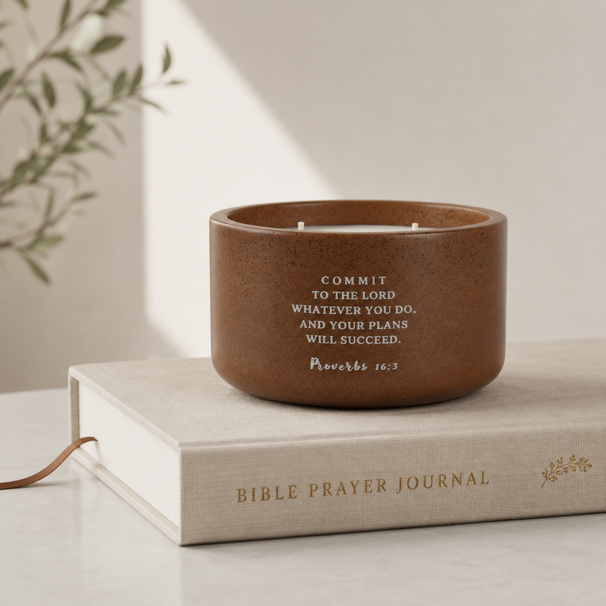 A brown concrete candle with white printed scripture text sits on top of a light beige Bible Prayer Journal book on a marble surface. A soft shadow and a blurred plant branch are visible in the background.