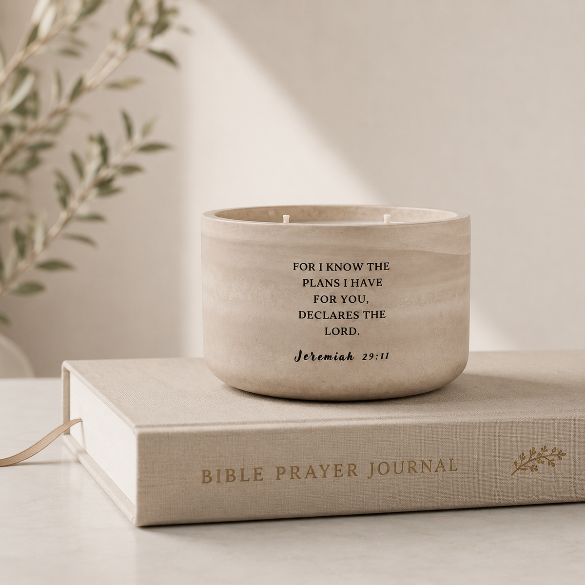 A small round candle in a light beige concrete holder sits on top of a hardcover book labeled Bible Prayer Journal. The candle has black printed text reading a Bible verse, and the scene is styled with soft natural light and blurred plant leaves in the background.