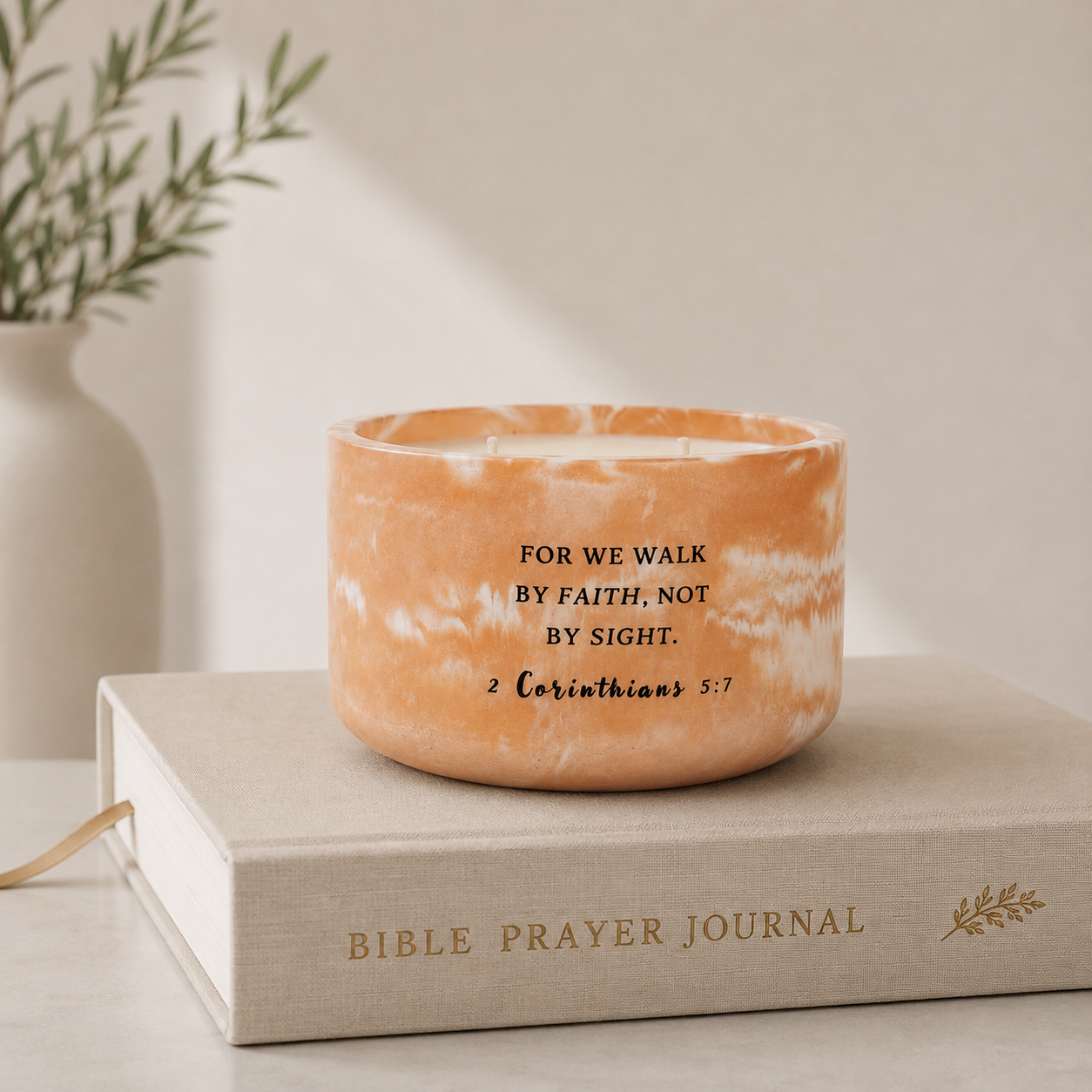 An orange marbled candle with black printed scripture text sits on top of a beige Bible Prayer Journal book, with a soft-focus background showing a white vase and greenery.