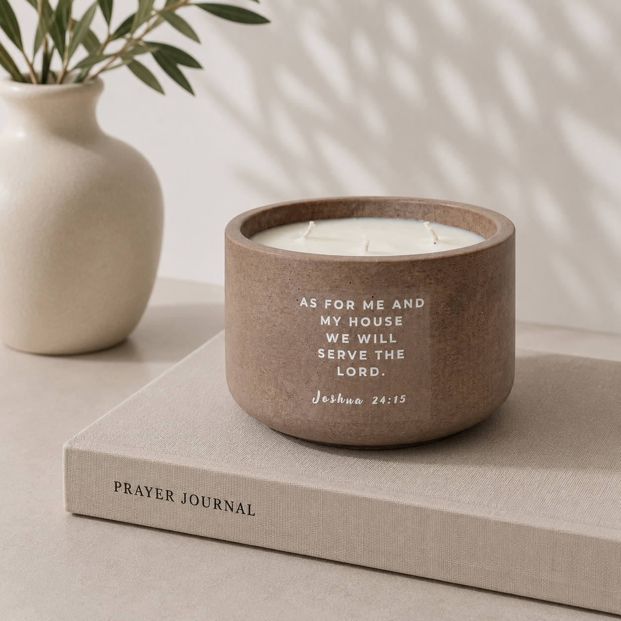 A round brown ceramic candle holder with white wax sits on a beige book labeled Prayer Journal. The candle holder has white text that reads, 'AS FOR ME AND MY HOUSE WE WILL SERVE THE LORD, Joshua 24:15