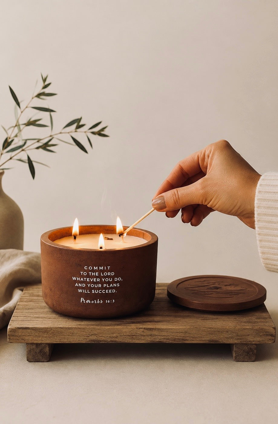 A wooden three-wick candle with a matching lid sits on a wooden tray, with a hand lighting it with a match. The candle has white text that reads 'COMMIT TO THE LORD, PRAY SO, AND YOUR PLANS WILL SUCCEED Psalm 37:5'.