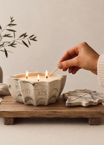 A hand uses a wooden match to light a three-wick candle in a sculptural, scalloped concrete holder engraved with the words 'simply BLESSED'. The candle sits on a rustic wooden tray next to its matching lid, with a soft-focus background of olive branches.
