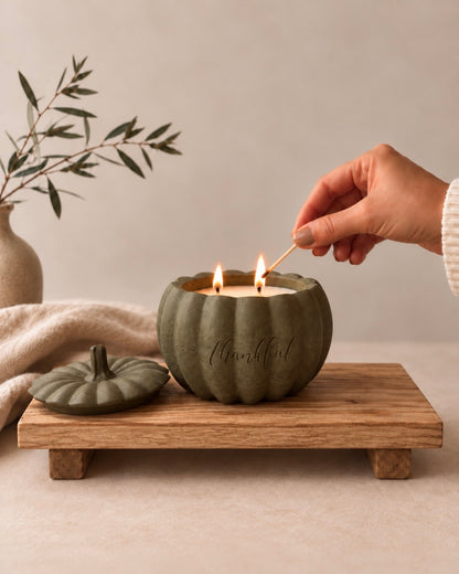 A hand holds a lit match to ignite a double-wick candle in a pumpkin-shaped ceramic holder. The candle holder is a muted sage green with the word 'thankful' embossed on the front.