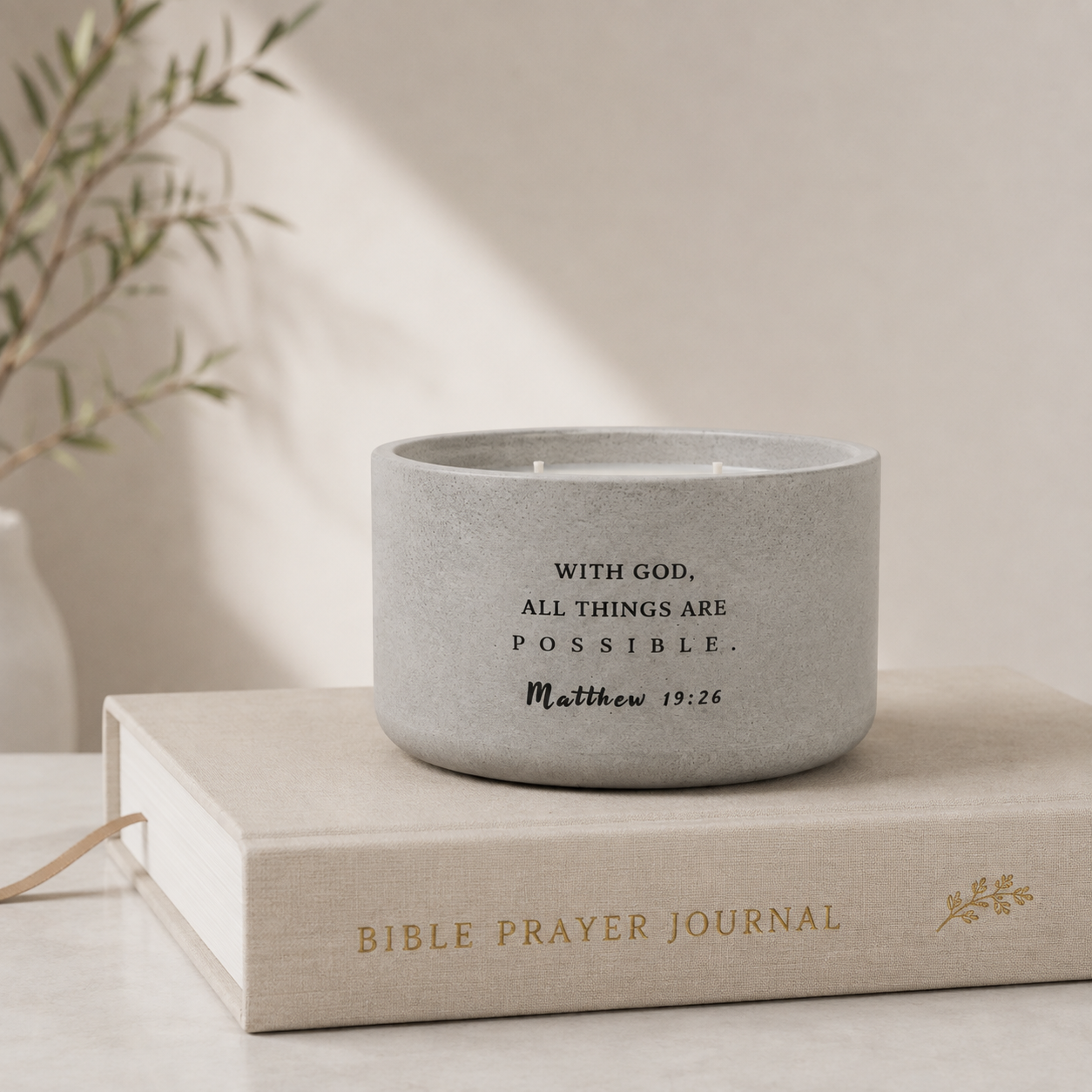 A concrete scripture candle with black printed scripture text sits on top of a beige Bible Prayer Journal book. The scene is softly lit with a warm neutral background and blurred greenery in the corner.