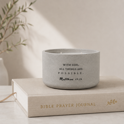 A concrete scripture candle with black printed scripture text sits on top of a beige Bible Prayer Journal book. The scene is softly lit with a warm neutral background and blurred greenery in the corner.