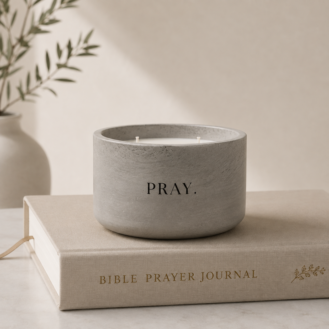 A gray concrete-textured candle jar with the word 'PRAY.' printed on the front, resting on top of a beige Bible Prayer Journal book.