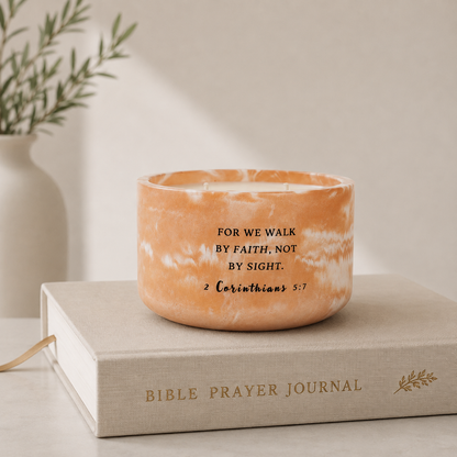 An orange marbled candle with black printed scripture text sits on top of a beige Bible Prayer Journal book, with a soft-focus background showing a white vase and greenery.