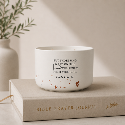 A white ceramic candle with black printed scripture text sits on top of a beige Bible Prayer Journal book, with a soft-focus vase and greenery in the background.