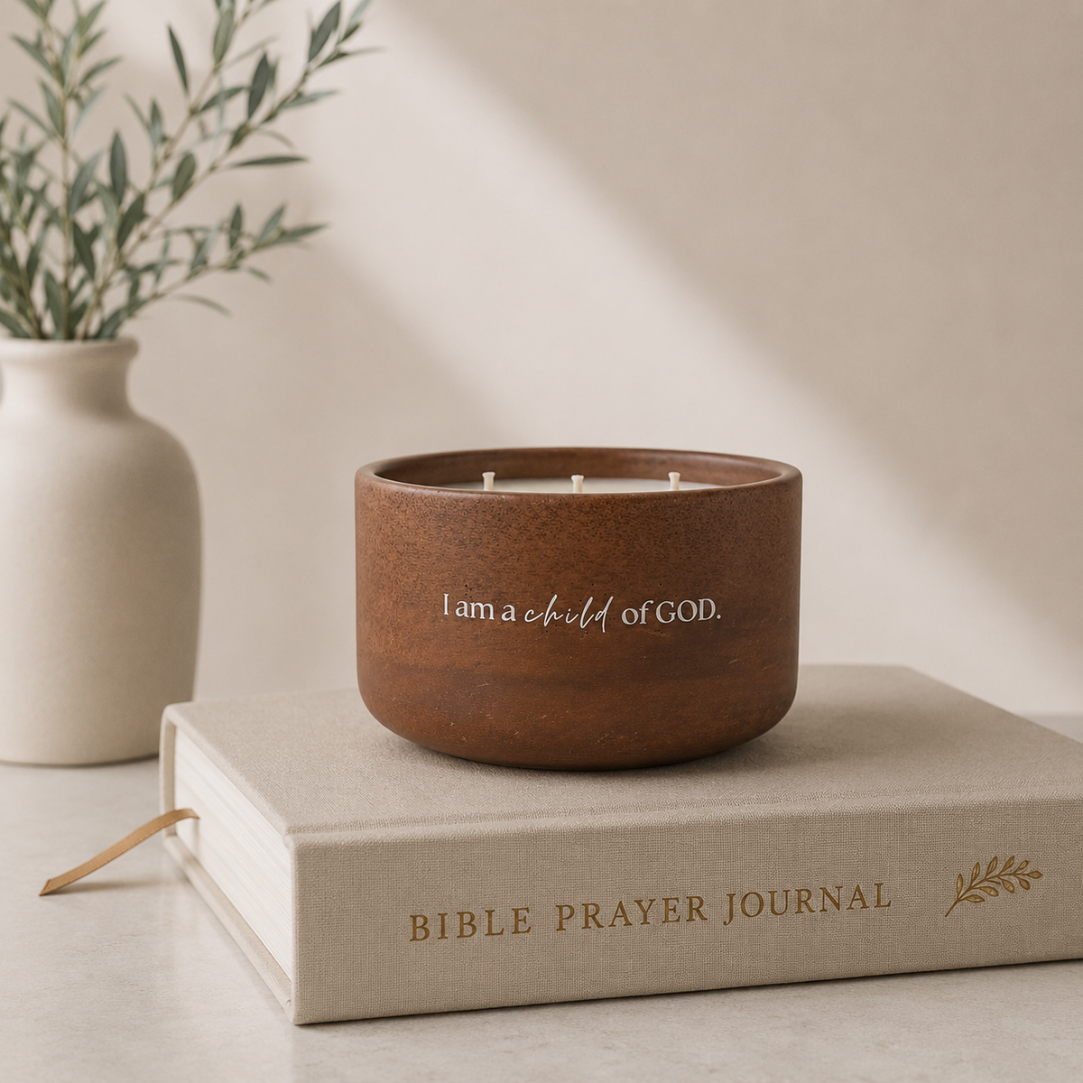 A brown wooden candle holder with white wicks and the text 'I am a child of GOD.' sits on top of a beige Bible Prayer Journal book, with a small vase of greenery in the background.