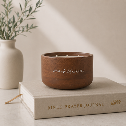 A brown wooden candle holder with white wicks and the text 'I am a child of GOD.' sits on top of a beige Bible Prayer Journal book, with a small vase of greenery in the background.