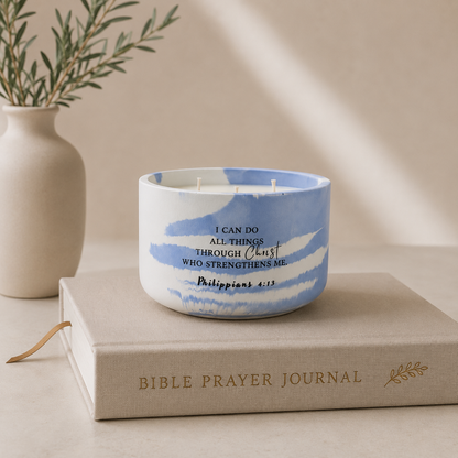 A white ceramic candle jar with blue watercolor-style brushstrokes and printed Bible verse text, styled on top of a beige Bible Prayer Journal book with a small olive branch vase in the background.