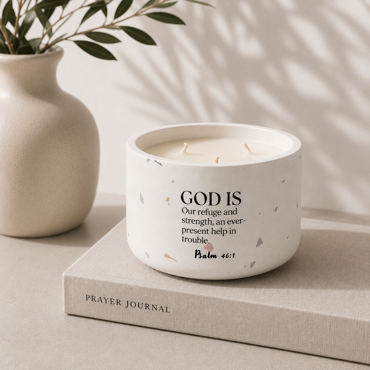 A white ceramic candle jar with a terrazzo-style speckled pattern sits on a beige book. The candle features black text that reads 'GOD IS Our refuge and strength, an ever-present help in trouble, Psalm 46:1'.