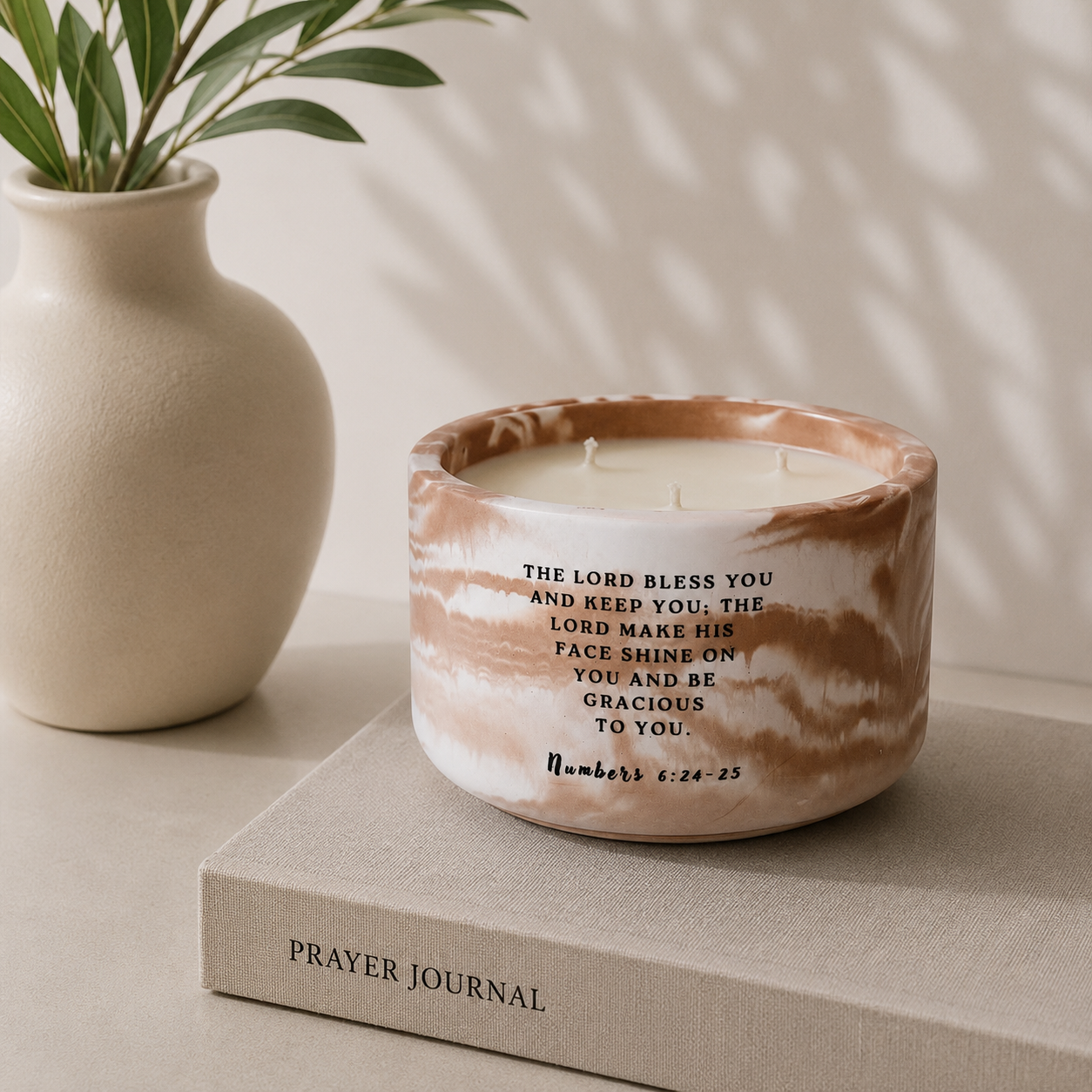 A ceramic candle in a round bowl with a brown and beige marbled pattern and black printed scripture text, Numbers 6:24-25 styled on a beige book next to a white vase with green leaves.