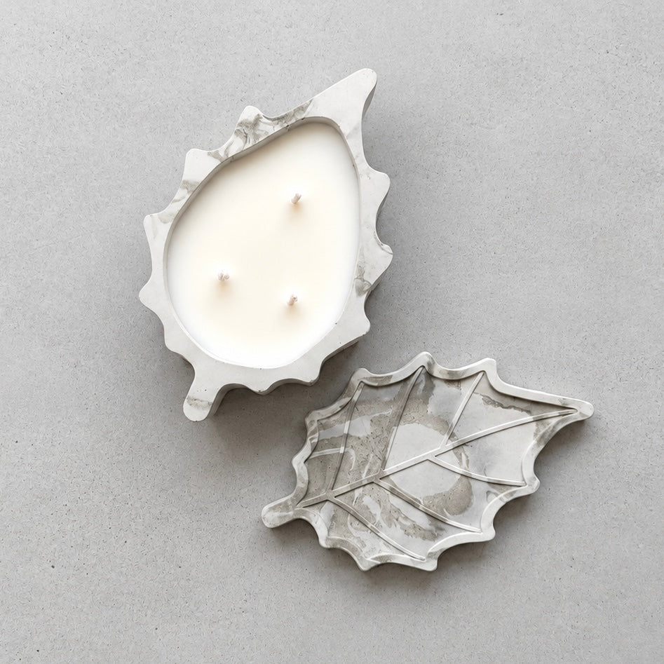 Overhead view of the leaf-shaped concrete candle with three unlit wicks and its matching marbled leaf lid beside it.