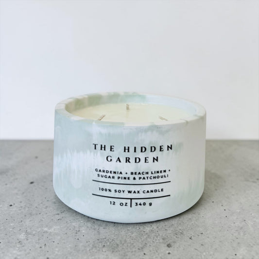 The Hidden Garden soy candle in marbled green and white concrete vessel with gardenia and beach linen scent on gray surface