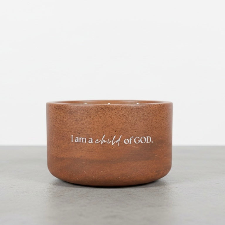 A small brown ceramic candle holder with the text 'I am a child of GOD.' printed in white on the front, set on a gray surface against a white background.